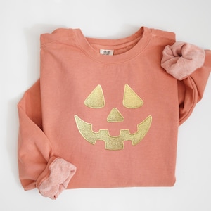 May include: A coral-colored sweatshirt with a gold, embroidered jack-o'-lantern face. The long-sleeved top has a crew neck and a matching scrunchie. The design is centered on the front of the shirt, perfect for Halloween.