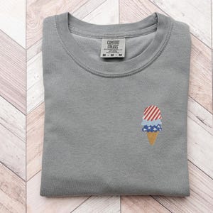 May include: A gray Comfort Colors sweatshirt with a small embroidered ice cream cone design. The ice cream cone features red and white stripes, blue stars, and a tan cone. The tag reads "Comfort Colors" and "M-M-M".