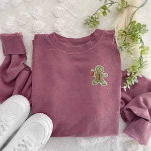 May include: A mauve-colored sweatshirt with a small embroidered gingerbread man holding a cup. The sweatshirt is paired with white sneakers and a sprig of greenery. The cuffs of the sleeves are rolled up.