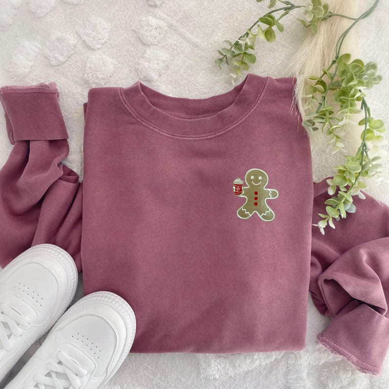 May include: A mauve-colored sweatshirt with a small embroidered gingerbread man holding a cup. The sweatshirt is paired with white sneakers and a sprig of greenery. The cuffs of the sleeves are rolled up.