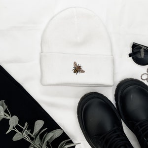 May include: A white beanie with a small embroidered bee design. The bee is brown, black, and yellow. The beanie is folded at the brim. Black boots, sunglasses, and a black garment are also in the image.