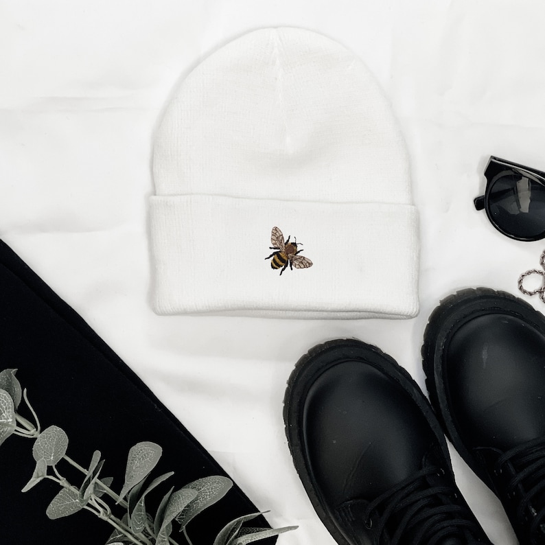 May include: A white beanie with a small embroidered bee design. The bee is brown, black, and yellow. The beanie is folded at the brim. Black boots, sunglasses, and a black garment are also in the image.