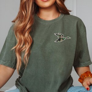 May include: A green short-sleeved t-shirt with a camouflage duck graphic on the front.