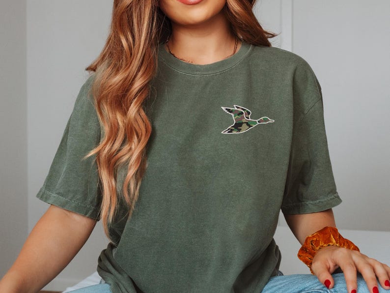May include: A green short-sleeved t-shirt with a camouflage duck graphic on the front.