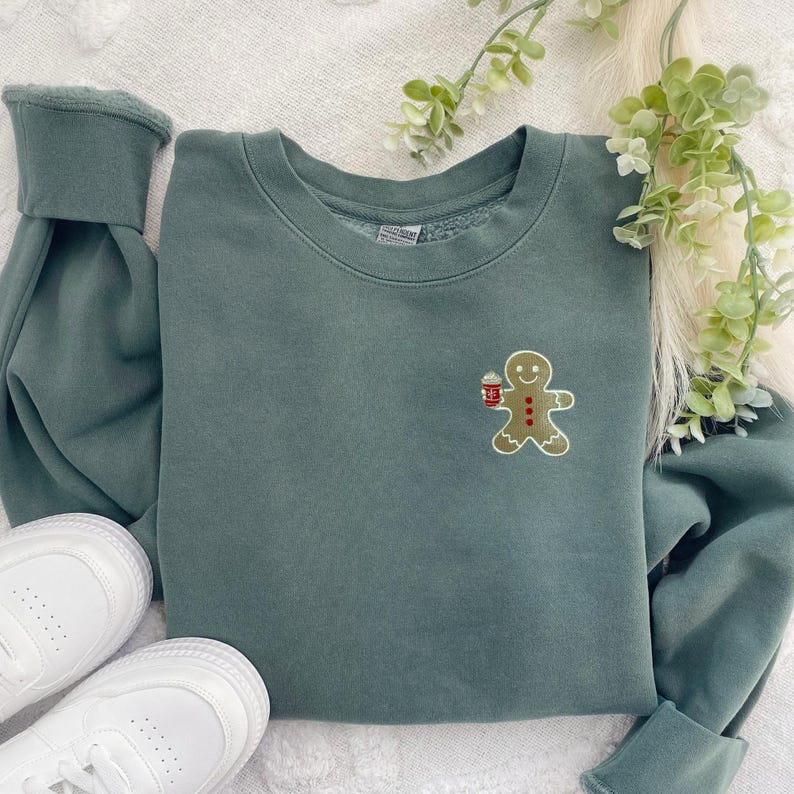 May include: A sage green sweatshirt featuring an embroidered gingerbread man holding a cup. The gingerbread man has red and brown accents. White sneakers are visible at the bottom of the image. The sweatshirt is a crew neck style.