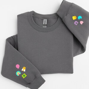 Peut inclure: Un sweat-shirt gris anthracite &agrave; col rond et manches longues. Le sweat-shirt a une petite &eacute;tiquette blanche sur le devant qui dit "Gildan" et une petite &eacute;tiquette blanche &agrave; l'int&eacute;rieur de la manche gauche qui dit "Softstyle". La manche droite a un graphique color&eacute; d'un arc-en-ciel, d'une &eacute;toile, d'un tr&egrave;fle et d'un c&oelig;ur. La manche gauche a un graphique color&eacute; d'un arc-en-ciel, d'une &eacute;toile, d'un tr&egrave;fle et d'un ballon.