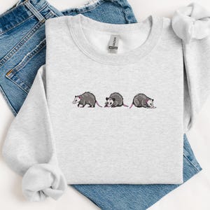 May include: A gray sweatshirt with a graphic of three opossums in a line. The opossums are gray with pink ears and noses.