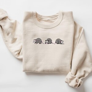 May include: A beige sweatshirt with a graphic of three opossums walking in a line. The opossums are embroidered in gray and pink.