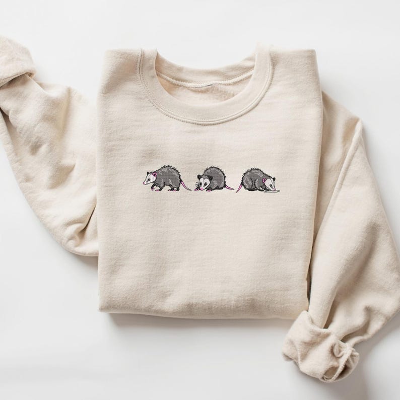 May include: A beige sweatshirt with a graphic of three opossums walking in a line. The opossums are embroidered in gray and pink.