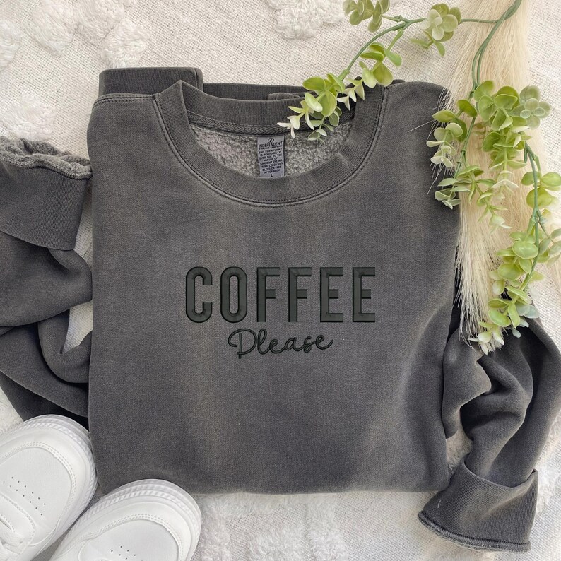 May include: A dark grey sweatshirt with the text "COFFEE Please" in black lettering. The sweatshirt is made of a soft, comfortable material and has a relaxed fit.