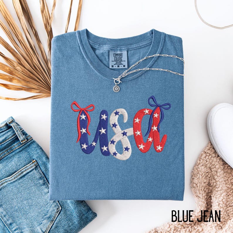 Pu&ograve; includere: T-shirt blu jeans con ricamo "USA" in rosso, bianco e blu. Le lettere sono decorate con stelle e fiocchi. Una collana d'argento con un ciondolo &egrave; appoggiata sulla maglietta. La maglietta &egrave; piegata su una superficie bianca.