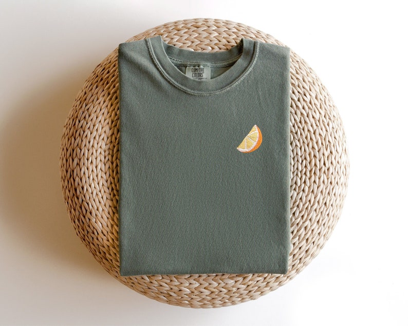 Puede incluir: Una camiseta verde con una peque&ntilde;a rodaja de naranja bordada en la parte delantera. La camiseta est&aacute; doblada y colocada sobre una estera de paja tejida.