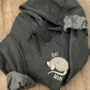 Puede incluir: Una sudadera con capucha gris oscuro con una rata bordada en blanco y el texto "Rat Mom".