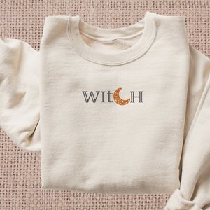 Puede incluir: Una sudadera de color crema con la palabra "WITCH" en texto negro y una luna creciente con un patrón marrón y naranja en el centro de la "I".