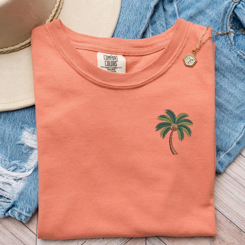 Op de afbeelding: Een koraaloranje T-shirt met een klein groen palmboommotief. Het T-shirt heeft een ronde hals en is gemaakt door Comfort Colors.