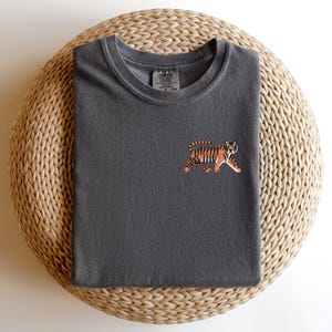May include: A dark gray t-shirt with a small embroidered tiger design on the front. The tiger is orange and black with a white background.