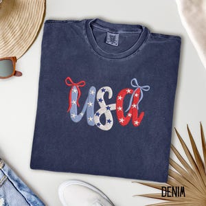 Pu&ograve; includere: Una maglietta blu navy con la scritta "USA" ricamata in rosso, bianco e blu. Le lettere sono decorate con stelle e fiocchi, evocando un tema patriottico. La maglietta &egrave; realizzata in un materiale morbido.