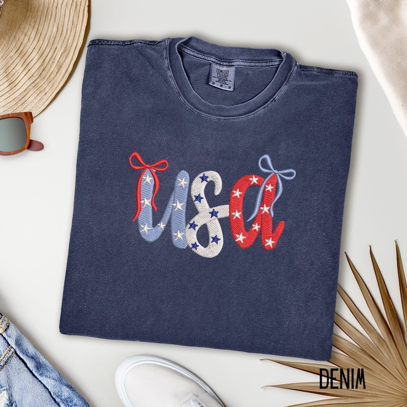 Pu&ograve; includere: Una maglietta blu navy con la scritta "USA" ricamata in rosso, bianco e blu. Le lettere sono decorate con stelle e fiocchi, evocando un tema patriottico. La maglietta &egrave; realizzata in un materiale morbido.