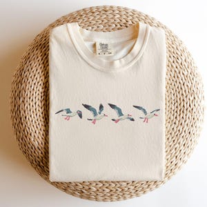 Puede incluir: Una camiseta de color crema con un dise&ntilde;o bordado de cinco gaviotas volando en l&iacute;nea. La camiseta tiene una peque&ntilde;a etiqueta que dice "Cotton Colors" y "S - P - CH".