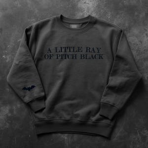 May include: Dark gray sweatshirt with the text "A LITTLE RAY OF PITCH BLACK" in dark gray. A small bat graphic is on the left sleeve. The sweatshirt has a crew neck and ribbed cuffs.