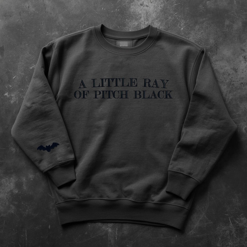 May include: Dark gray sweatshirt with the text "A LITTLE RAY OF PITCH BLACK" in dark gray. A small bat graphic is on the left sleeve. The sweatshirt has a crew neck and ribbed cuffs.