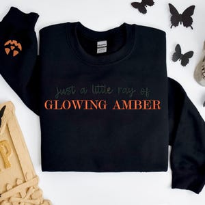 May include: Black sweatshirt with the text "just a little ray of GLOWING AMBER" in green and orange. The left sleeve has an orange pumpkin design. Black butterfly decorations are scattered around the sweatshirt.