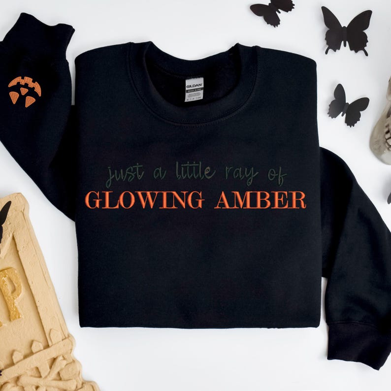 May include: Black sweatshirt with the text "just a little ray of GLOWING AMBER" in green and orange. The left sleeve has an orange pumpkin design. Black butterfly decorations are scattered around the sweatshirt.