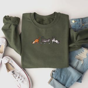 May include: A green sweatshirt with three embroidered cats in a row. The cats are walking in different directions. The sweatshirt is laid flat on a white surface.