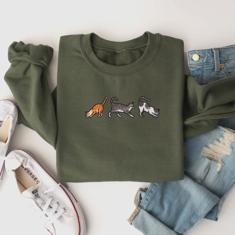 May include: A green sweatshirt with three embroidered cats in a row. The cats are walking in different directions. The sweatshirt is laid flat on a white surface.