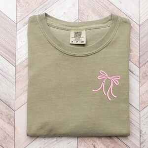 以下が含まれることがあります： 前面にピンクのリボンが刺繍された緑色のTシャツ。クルーネックと半袖。シャツのタグには「Comfort Colors」と書いてあります。
