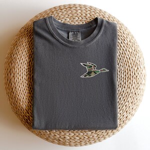 May include: A dark gray t-shirt with a camouflage duck design embroidered on the front. The shirt has a crew neck and short sleeves.