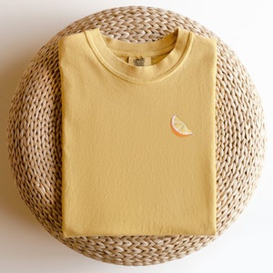 Puede incluir: Una camiseta amarilla mostaza con una peque&ntilde;a rodaja de naranja bordada en la parte delantera. La camiseta est&aacute; doblada y colocada sobre una estera de paja tejida.