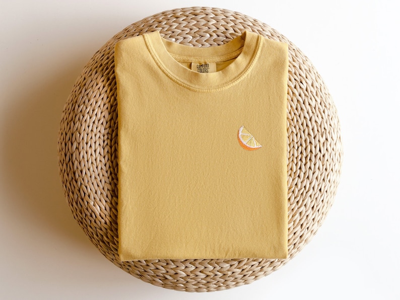 Puede incluir: Una camiseta amarilla mostaza con una peque&ntilde;a rodaja de naranja bordada en la parte delantera. La camiseta est&aacute; doblada y colocada sobre una estera de paja tejida.