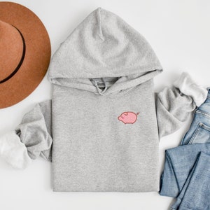 K&ouml;nnte beinhalten: Ein grauer Kapuzenpullover mit einem kleinen rosa Schweinchen, das auf der Vorderseite gestickt ist.