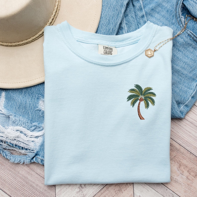 Op de afbeelding: Een lichtblauw t-shirt met een palmboom-print en de tekst "Comfort Colors" op het label.