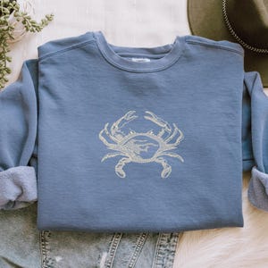K&ouml;nnte beinhalten: Ein blaues Sweatshirt mit einem hellbeigen Krabbenmuster, das auf der Vorderseite gestickt ist. Das Sweatshirt hat einen Rundhalsausschnitt und lange &Auml;rmel mit umgeschlagenen Manschetten. Das Krabbenmuster ist mittig auf der Brust platziert.