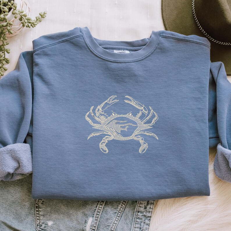K&ouml;nnte beinhalten: Ein blaues Sweatshirt mit einem hellbeigen Krabbenmuster, das auf der Vorderseite gestickt ist. Das Sweatshirt hat einen Rundhalsausschnitt und lange &Auml;rmel mit umgeschlagenen Manschetten. Das Krabbenmuster ist mittig auf der Brust platziert.