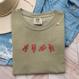 May include: A green t-shirt with a red lobster design. The shirt has the text "COMFORT COLORS" on the tag. The shirt is folded and laid on a wooden surface.