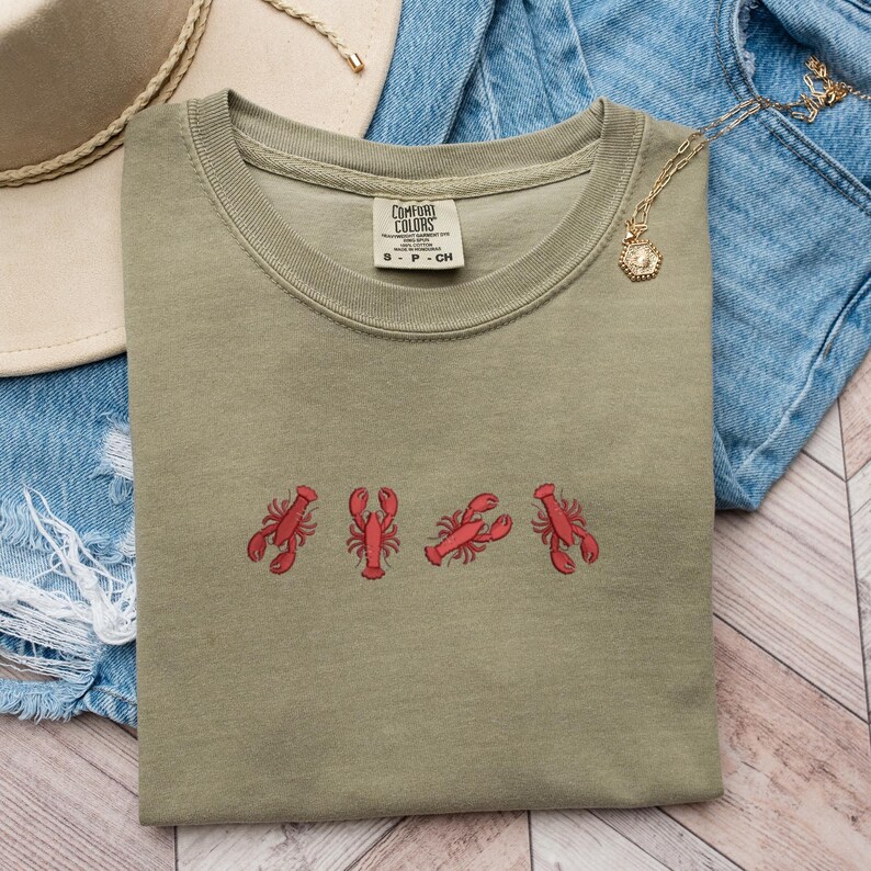 May include: A green t-shirt with a red lobster design. The shirt has the text "COMFORT COLORS" on the tag. The shirt is folded and laid on a wooden surface.