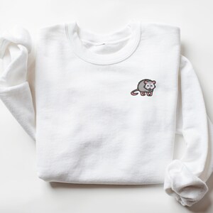 May include: White sweatshirt with a small embroidered image of a gray opossum with pink ears and nose.