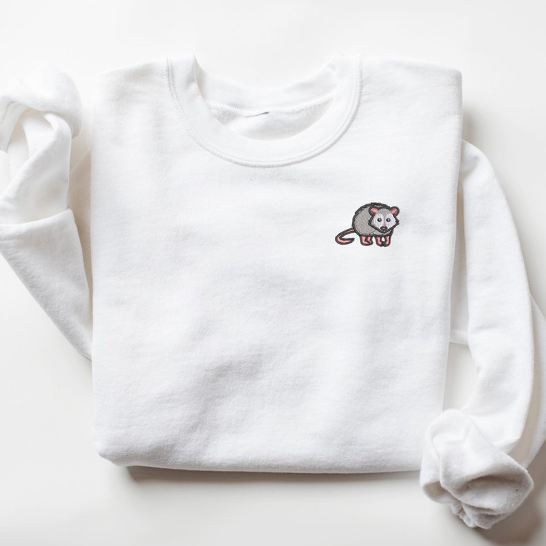 May include: White sweatshirt with a small embroidered image of a gray opossum with pink ears and nose.
