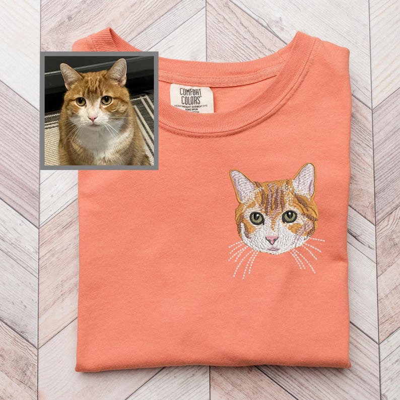 May include: Coral-colored t-shirt with an embroidered orange tabby cat face on the front. A small framed photo of a similar cat is in the upper left corner. The shirt has a crew neck and a tag that reads "Comfort Colors".