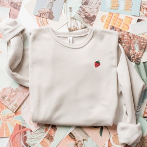 May include: A light beige sweatshirt with a small embroidered strawberry on the front. The sweatshirt is laying on a pile of vintage postcards.