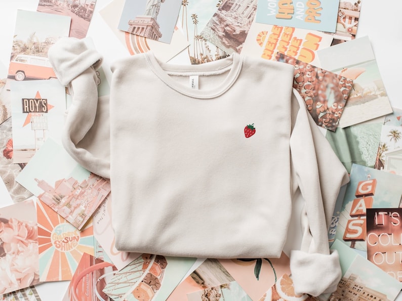 May include: A light beige sweatshirt with a small embroidered strawberry on the front. The sweatshirt is laying on a pile of vintage postcards.