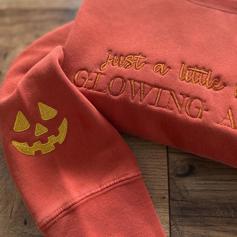May include: A coral-colored sweatshirt with embroidered text that reads "just a little glowing". The sleeve features a gold embroidered jack-o'-lantern face. The sweatshirt is folded on a wooden surface.