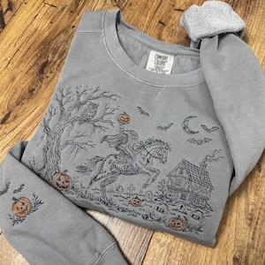 May include: A grey crewneck sweatshirt featuring a black embroidered scene of a headless horseman riding a horse, a tree with an owl, a house, tombstones, bats, a crescent moon, and orange pumpkins. The inner neck label reads COMFORT COLORS.