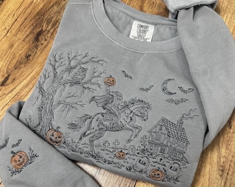 May include: A grey crewneck sweatshirt featuring a black embroidered scene of a headless horseman riding a horse, a tree with an owl, a house, tombstones, bats, a crescent moon, and orange pumpkins. The inner neck label reads COMFORT COLORS.