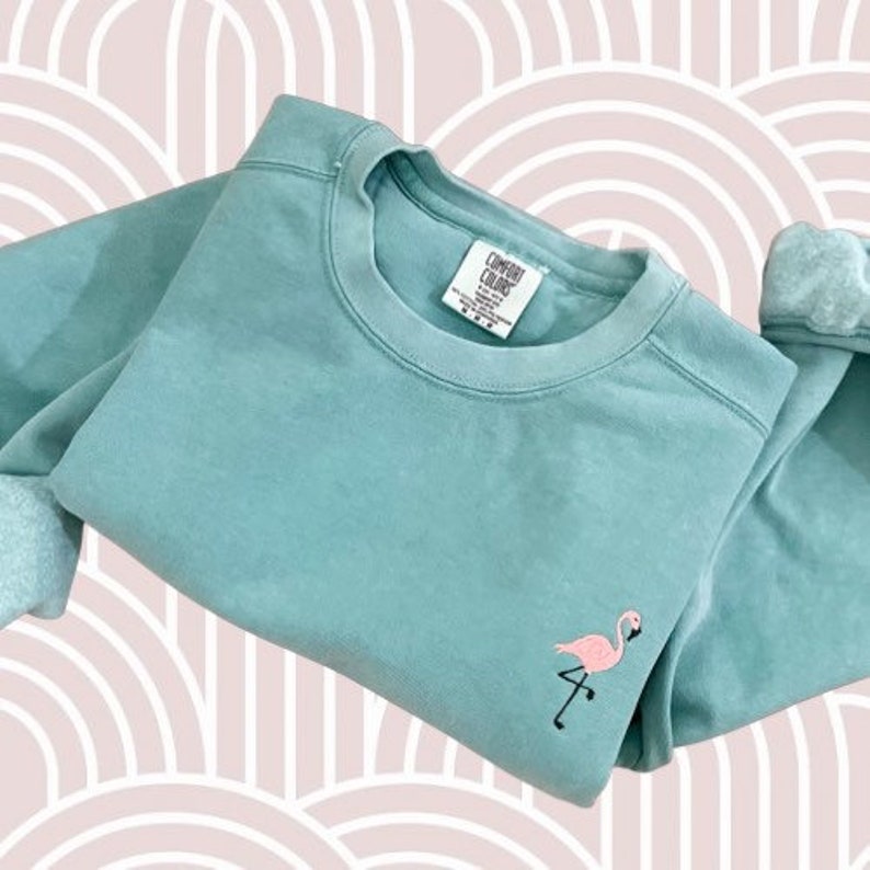 May include: A teal-colored sweatshirt with a pink flamingo embroidered on the front. The sweatshirt has a crew neck and long sleeves. The tag reads "Comfort Colors". The background is a pink and white pattern.
