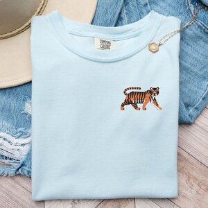 May include: A light blue t-shirt with a tiger graphic on the front. The t-shirt has a crew neck and short sleeves. The tiger is brown and orange with black stripes. The t-shirt is laying on a pair of blue jeans and a brown hat.