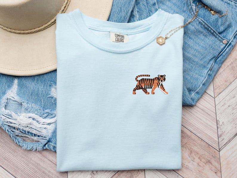 May include: A light blue t-shirt with a tiger graphic on the front. The t-shirt has a crew neck and short sleeves. The tiger is brown and orange with black stripes. The t-shirt is laying on a pair of blue jeans and a brown hat.